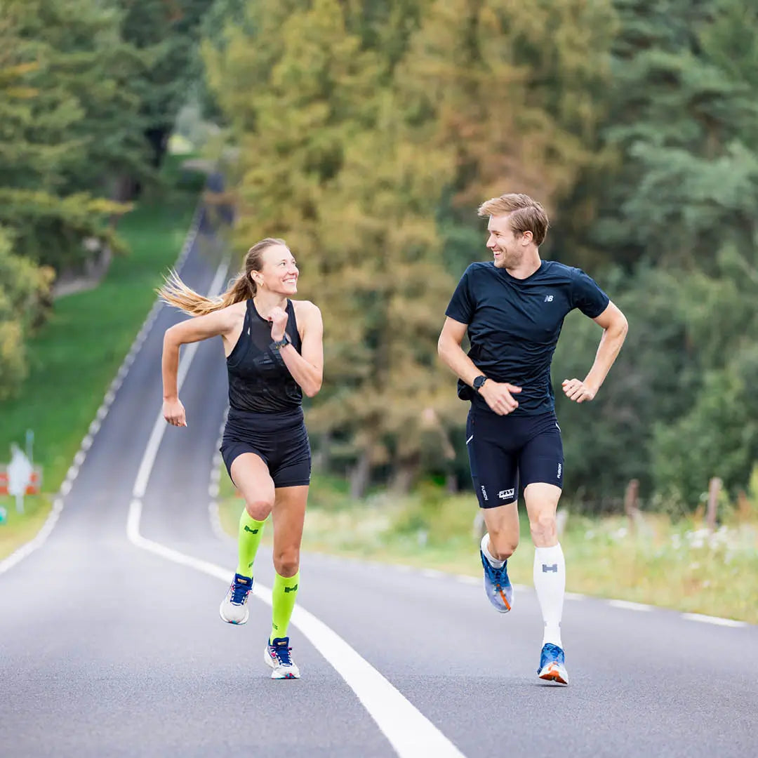 Man en vrouw hardlopen over de weg en dragen de gele en witte Pro Compressiekousen.