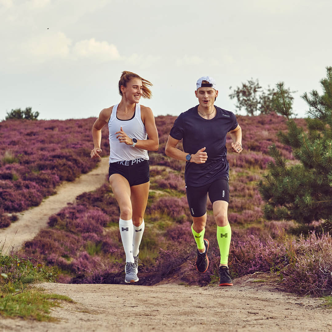 Man en vrouw hardlopen tussen paarse heide. De vrouw draagt witte compressiekousen en de man gele compressietubes