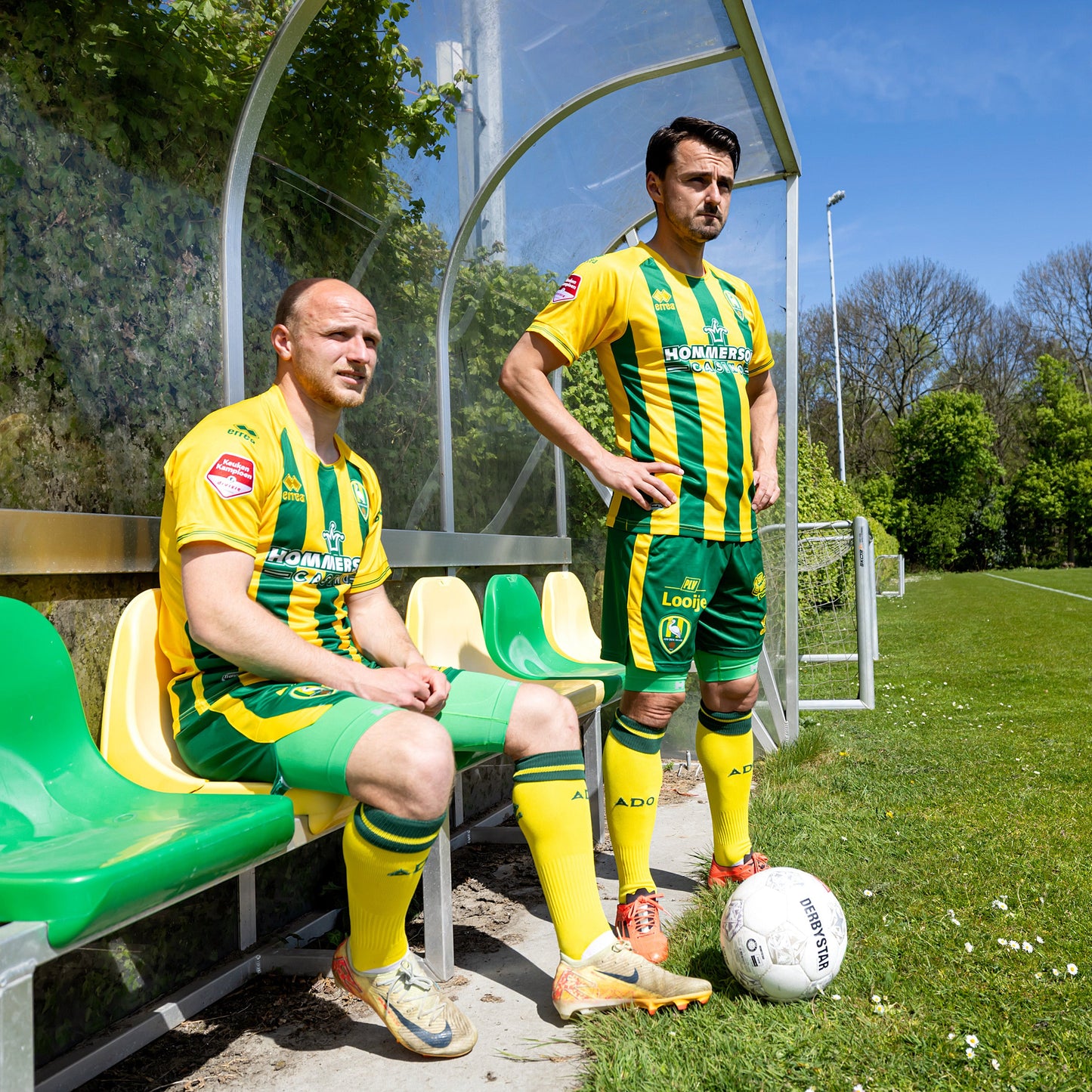 twee voetballers van ADO Den Haag in de dugout en kijken naar naar het grasveld